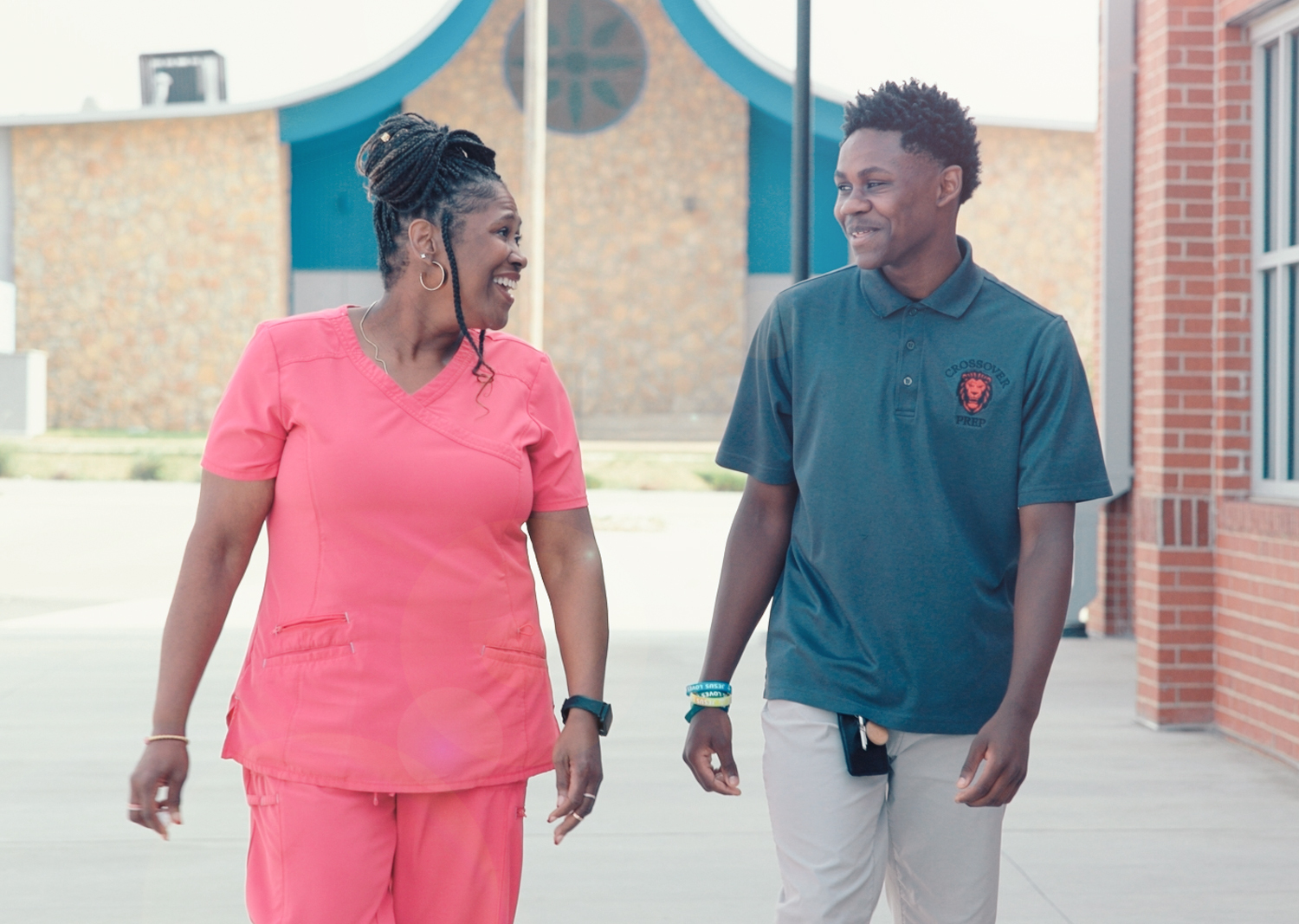 A smiling mother and her teenage son walk together outside a school building, laughing and looking at each other.