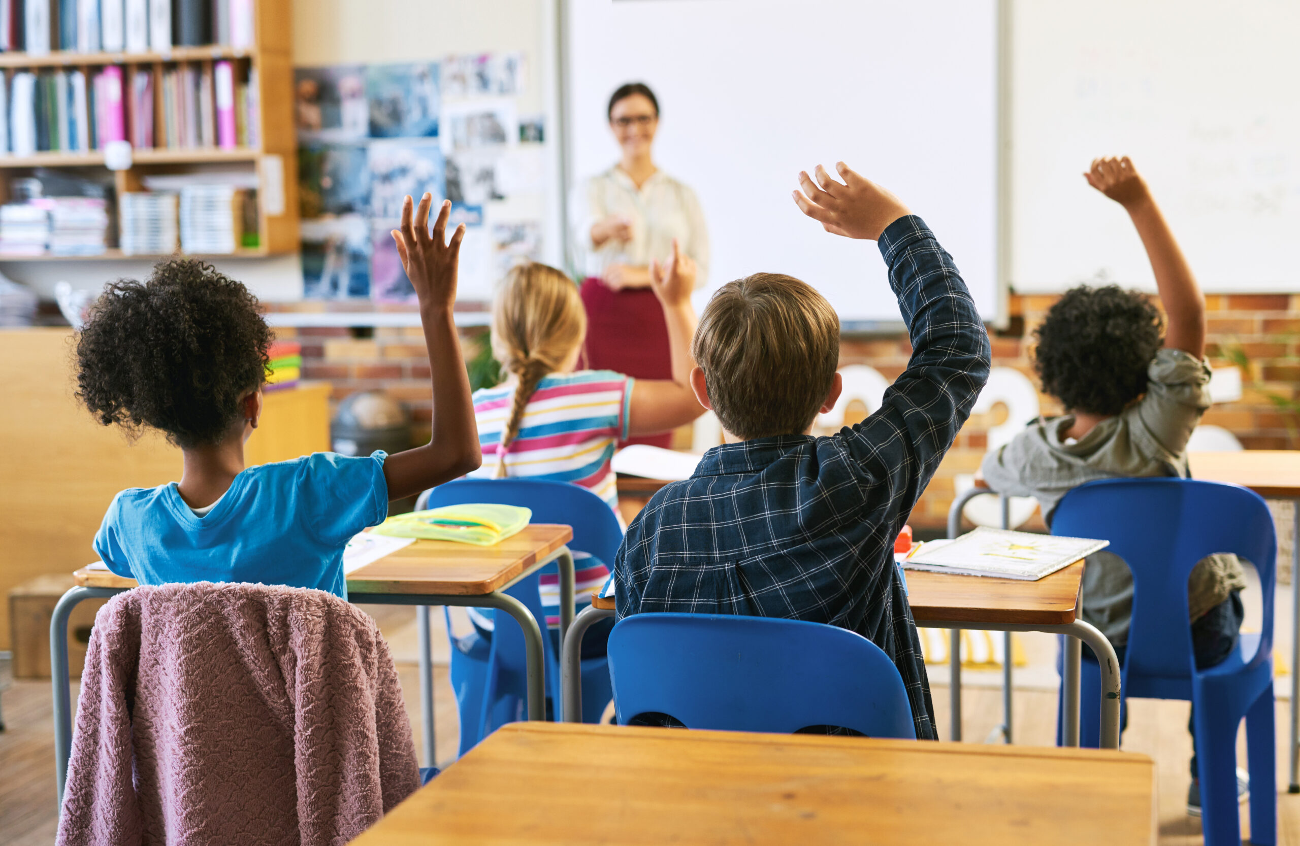 Students in a classroom raising their hands to answer a question while a teacher stands at the front