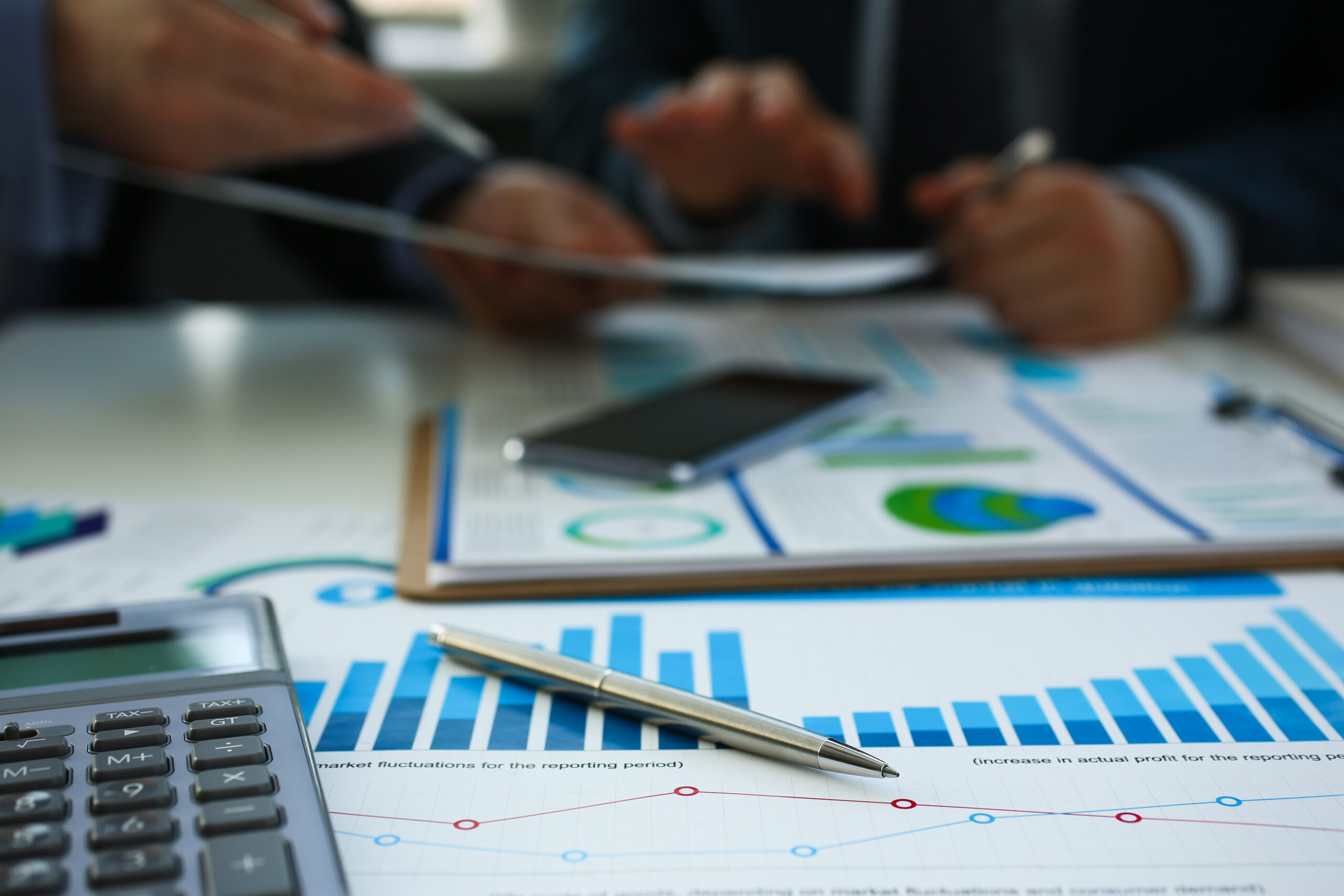 Close-up of financial charts and graphs on a desk with a calculator, pen, and smartphone while two people review documents in the background.