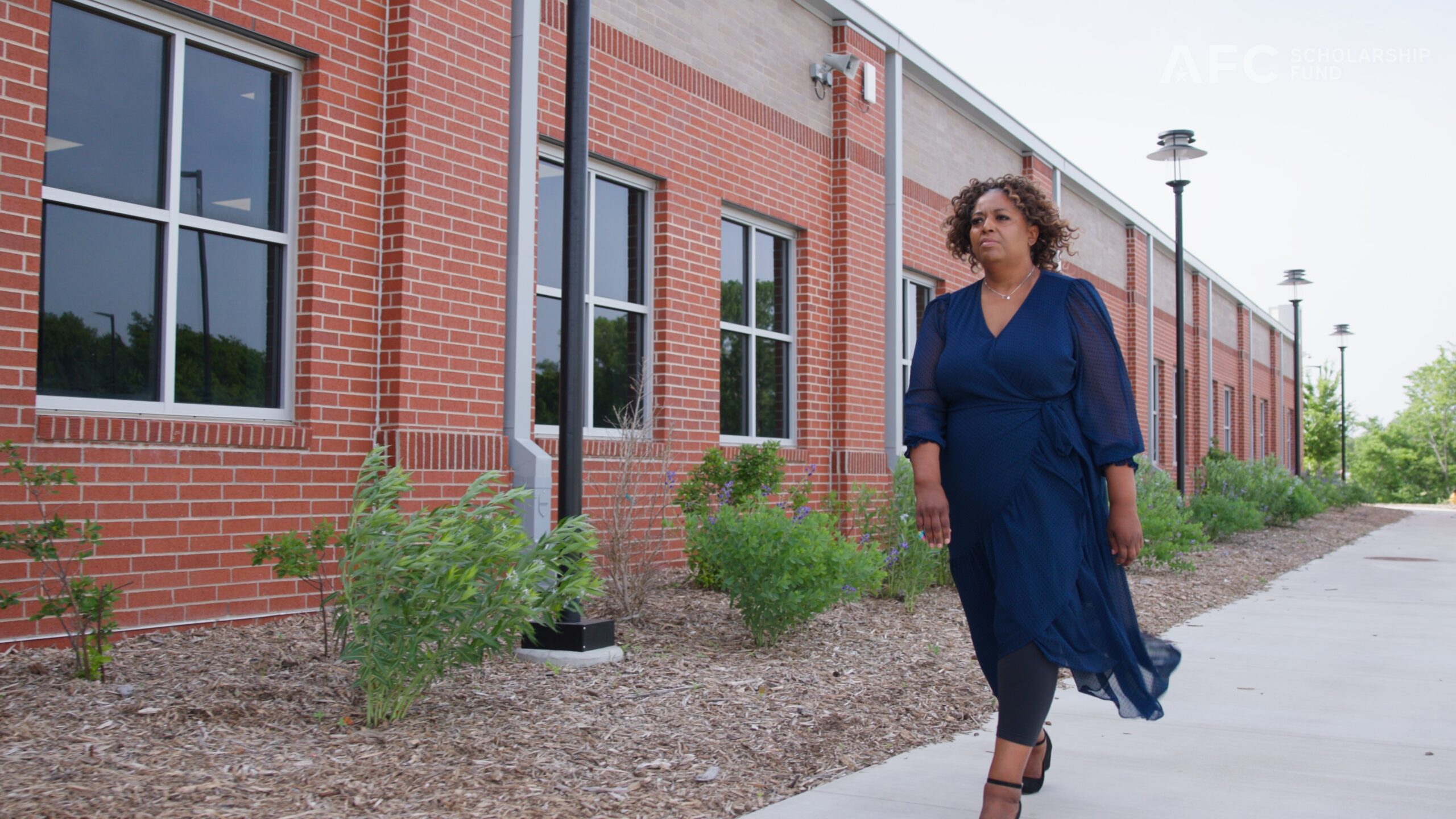 Woman walking outside a brick school building along a sidewalk lined with plants.