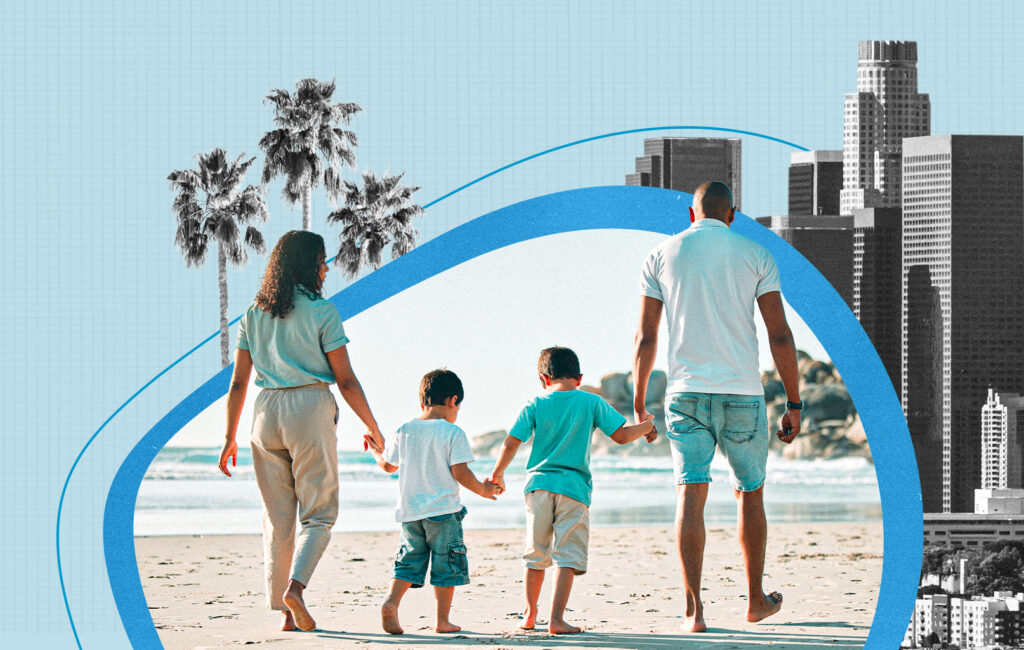 A family walks along a beach holding hands, with the Los Angeles skyline in the background.